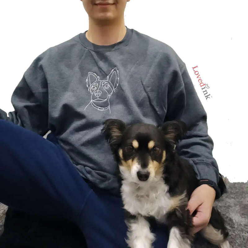 A person wearing a dark grey sweatshirt with a custom white line-art embroidery of their pet, while sitting with their small black, tan, and white dog. The image showcases the Lovedink logo and the high-quality resemblance of the custom design